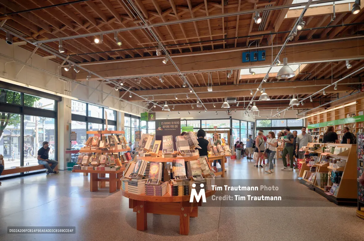 Inside Powell's City of Books in Portland's Pearl District, warm tungsten track lighting illuminates the famous bookstore's exposed timber ceiling and polished concrete floors. Circular wooden display tables showcase carefully curated book selections while customers browse leisurely among the towering shelves. Floor-to-ceiling windows bathe the space in natural daylight, revealing the bustling Burnside Street beyond and creating an inviting atmosphere where literature and community converge.