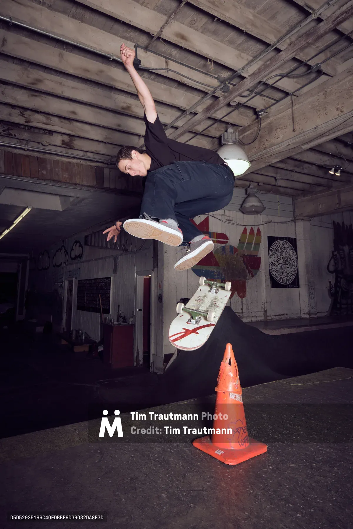 A skateboarder executes a dynamic jump over a weathered orange traffic cone inside Commonwealth Skateboarding in Portland's Buckman neighborhood. The dramatic low-angle perspective captures the athlete mid-flight against the industrial basement ceiling, with exposed beams and graffiti-adorned concrete walls creating a raw urban backdrop. The skater's concentrated expression and athletic form demonstrate the precision required for this technical maneuver. Harsh artificial lighting casts deep shadows across the underground skate space, emphasizing the gritty authenticity of Portland's skateboarding culture.