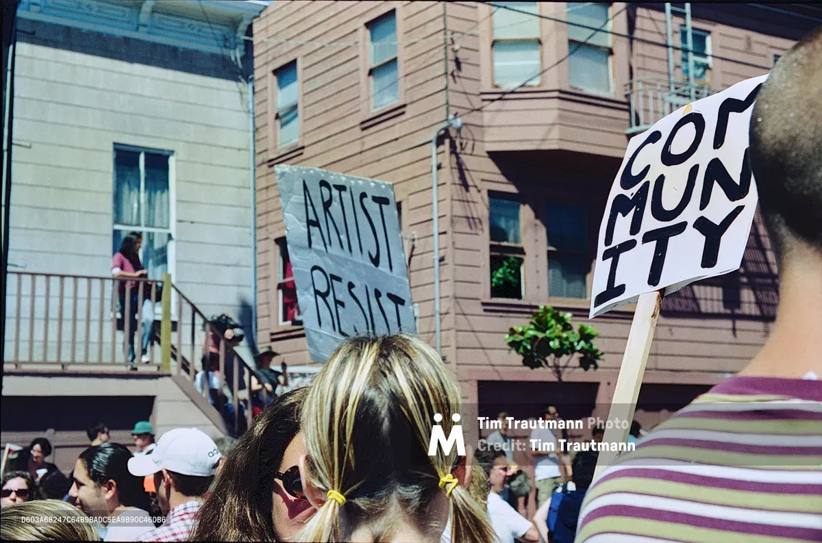In the heart of San Francisco's Mission District, protesters rally against displacement with handmade signs reading "ARTIST RESIST" and "COMMUNITY." The demonstration unfolds beneath characteristic wooden apartment buildings on Bryant Street, where residents of all ages gather in defiant solidarity. Golden afternoon light bathes the crowd, creating an intimate yet urgent atmosphere as longtime community members make their voices heard against the forces of gentrification.