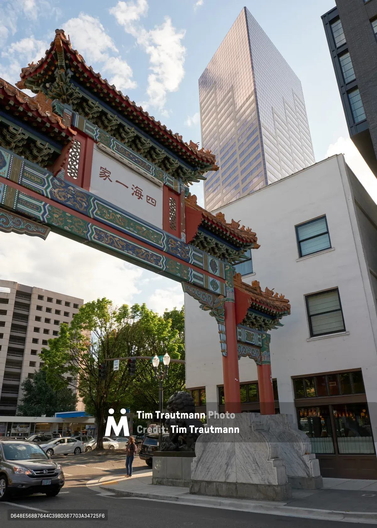 A striking architectural juxtaposition unfolds in Portland's Chinatown as an ornate Chinese ceremonial gate, adorned with vermillion lacquer and intricate dragon carvings, stands sentinel before gleaming glass towers. The traditional paifang, supported by granite guardian lions, creates a dramatic foreground against the contemporary urban backdrop of rose-tinted skyscrapers and residential buildings. Afternoon light casts gentle shadows across the busy street where pedestrians and vehicles navigate between centuries of architectural heritage.