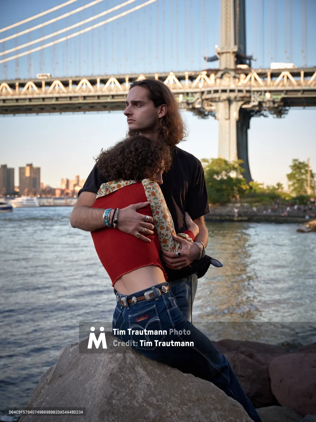 A tender moment unfolds along the East River waterfront as a young man with long dark hair cradles a woman with curly brown hair against the dramatic backdrop of the Manhattan Bridge. The couple sits on weathered rocks in Brooklyn Bridge Park, bathed in warm golden light that illuminates the bridge's suspension cables and stone towers. The woman wears a vibrant red ribbed top and floral shirt, while the man is dressed in a black t-shirt and vintage denim, their intimate embrace captured during the magic hour as the city skyline glows softly in the distance.