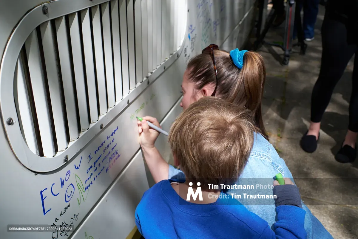 A TriMet train operator in a blue scrunchie and denim jacket leans over her young child in a royal blue sweatshirt as they write farewell messages on the white exterior of a MAX Type 1 train car during its retirement ceremony at Holladay Park in Portland's Lloyd District. Both concentrate intently as they add their handwritten notes to the growing collection of colorful marker messages covering the train's surface. The circular ventilation grate and industrial design of the vintage transit car frame their tender moment of personal connection to the retiring fleet.