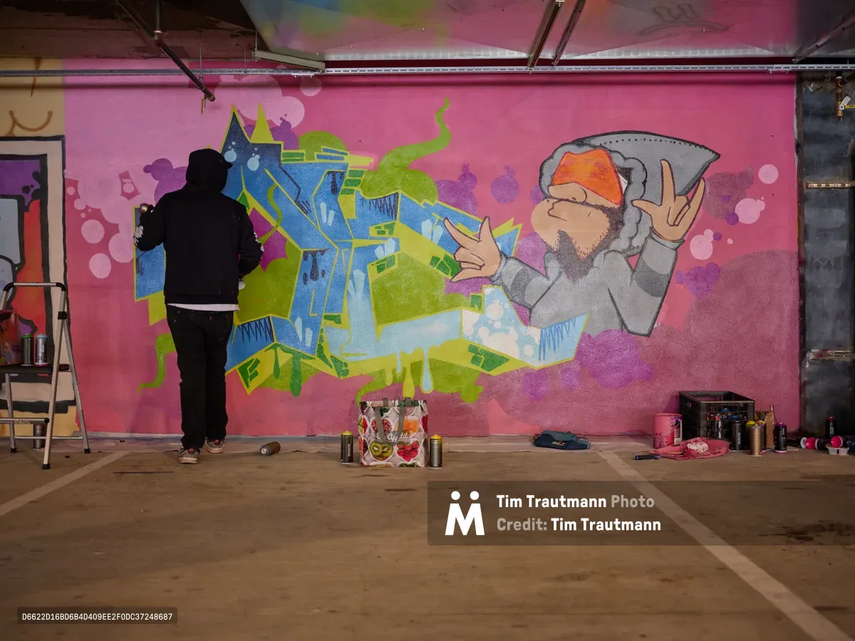 A silhouetted muralist works on an explosive burst of color against the magenta wall of Clay Creative's parking garage in Portland's Hosford-Abernethy neighborhood. The emerging artwork features fantastical architectural forms in electric blues and lime greens, punctuated by a bearded figure making peace signs, while spray cans and art supplies scatter across the concrete floor. Raw industrial ceiling elements frame this intimate glimpse into Southeast Portland's creative underground, where urban art transforms utilitarian spaces into canvases of imagination.