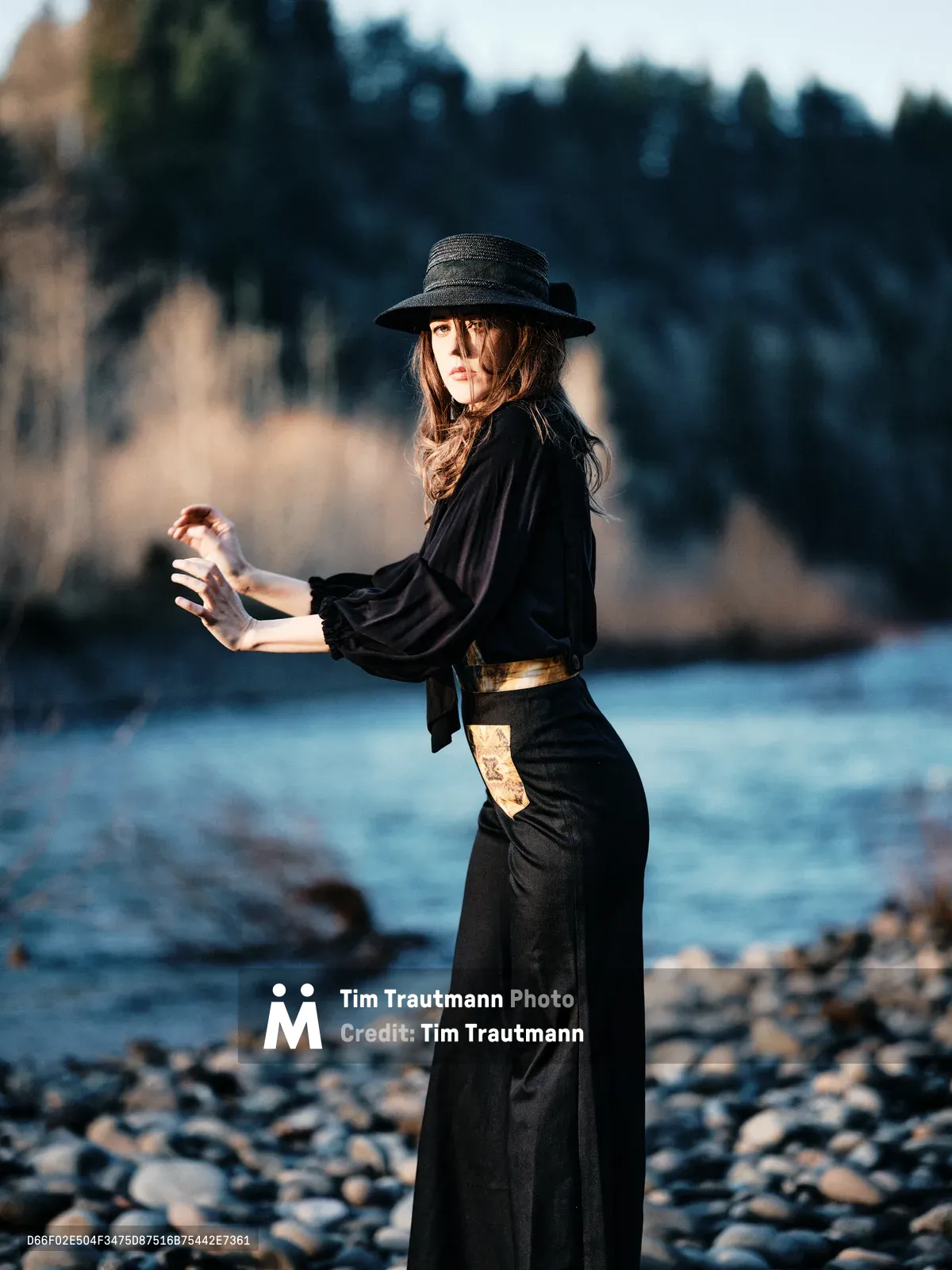 A woman in flowing black attire and wide-brimmed hat stands in contemplative pose along the rocky shoreline of the Columbia River Gorge. Golden hour light catches her profile as she cradles a glass bottle, creating an intimate moment against the backdrop of Oregon's forested riverbank. The composition balances her silhouetted form against the soft blur of water and distant treeline, evoking themes of solitude and reflection in nature.