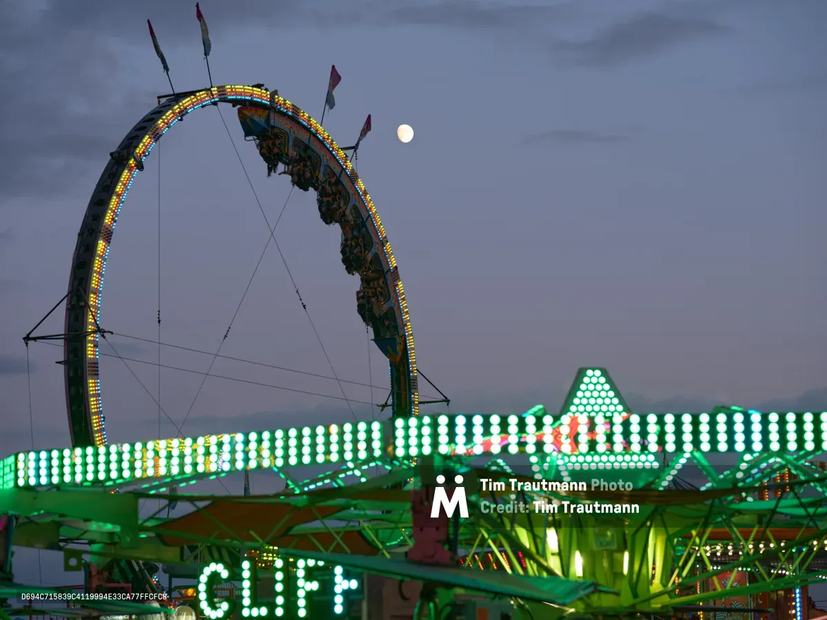Ornate carnival ride architecture dominates the frame against a dusky twilight sky at the Clark County Fair in Vancouver, Washington. A sweeping arc of multicolored LED lights traces an elaborate ride structure while a pale moon hovers in the purple-gray evening sky. Below, the geometric patterns of ride lighting create a constellation of green and white bulbs, with the word 'CLIFF' prominently illuminated in bold carnival lettering. The scene captures the magical transition from day to night when fair lights begin their nightly spectacle.