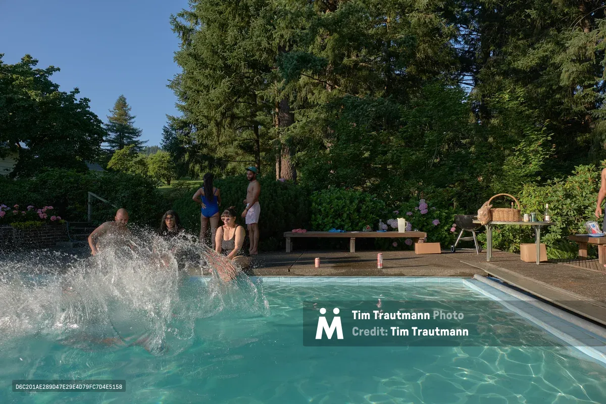 A dynamic summer scene unfolds at a private pool in Newberg, Oregon's wine country, where a man creates a dramatic splash as he dives into turquoise water. Family and friends gather around the pool deck under towering evergreens, with some relaxing poolside while others stand ready to join the aquatic fun. The late afternoon light filters through the lush canopy, illuminating picnic supplies and creating a perfect backdrop of domestic leisure in the Pacific Northwest. The scene captures the essence of summer hospitality against Oregon's natural splendor.