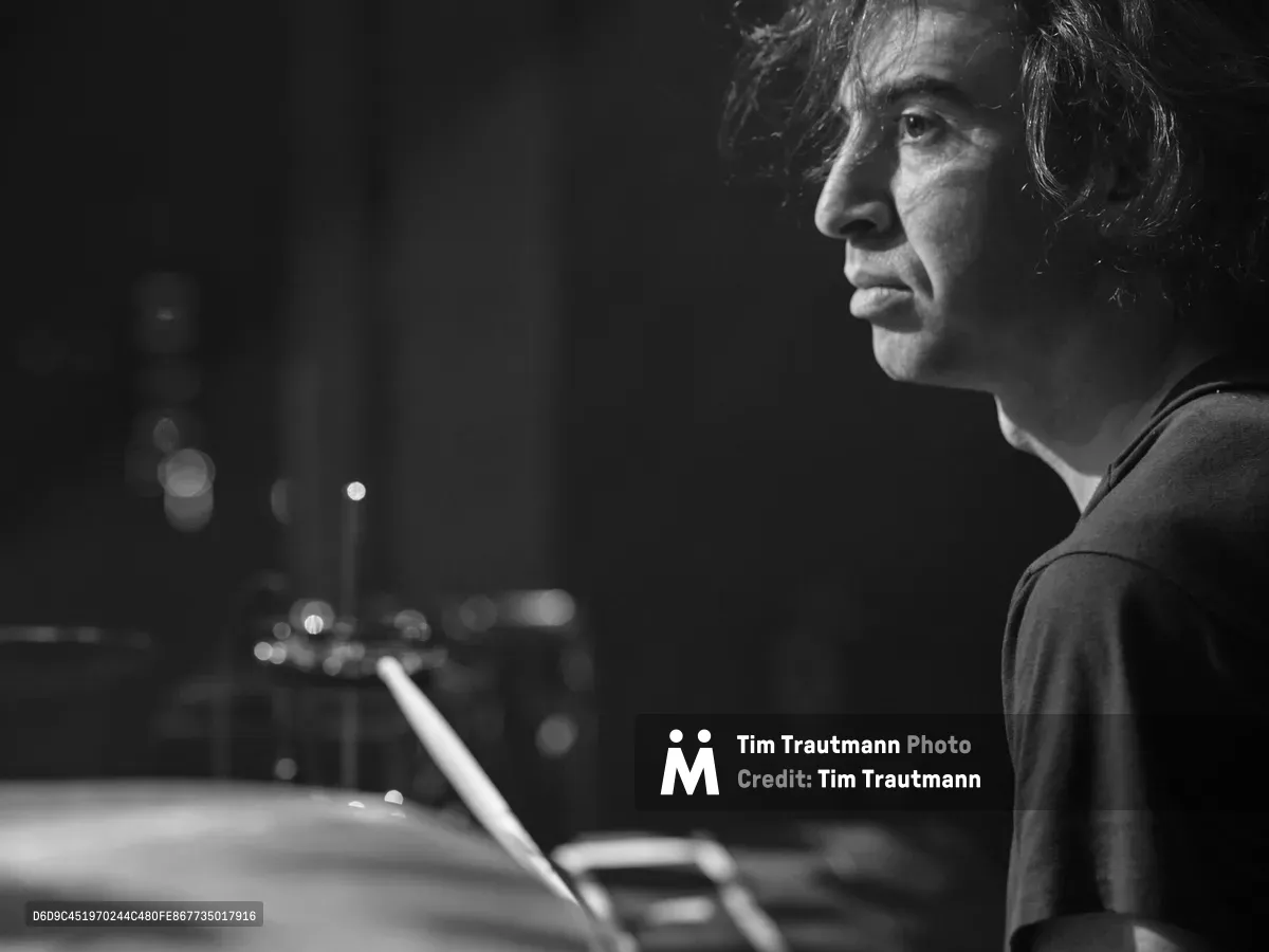 Julian Dorio of Band of Skulls sits in quiet contemplation behind his drum kit at the legendary Webster Hall in Manhattan during the band's 10 Year Anniversary Tour. The black and white portrait captures the drummer in profile against a backdrop of soft stage lights, with drumsticks poised in his hands. The atmospheric lighting creates dramatic shadows across his face while the blurred venue lights in the background suggest the intimate energy of this historic East Village music venue.