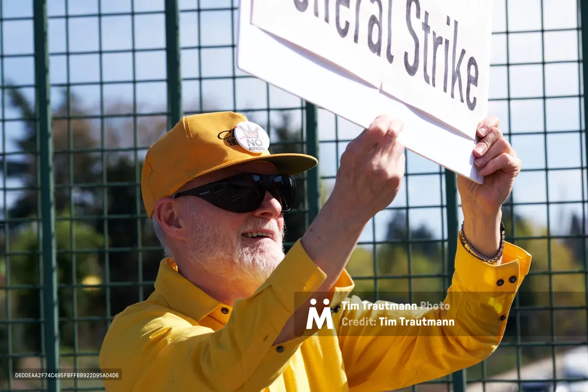 A determined activist in a bright yellow shirt and baseball cap holds aloft a white banner reading "General Strike" during a protest action near North Portland's Interstate 5 overpass. The man's weathered face shows quiet resolve as golden afternoon sunlight illuminates the scene, while chain-link fencing and urban infrastructure frame the background. His raised arms and focused expression capture the earnest spirit of grassroots political organizing in the Pacific Northwest.