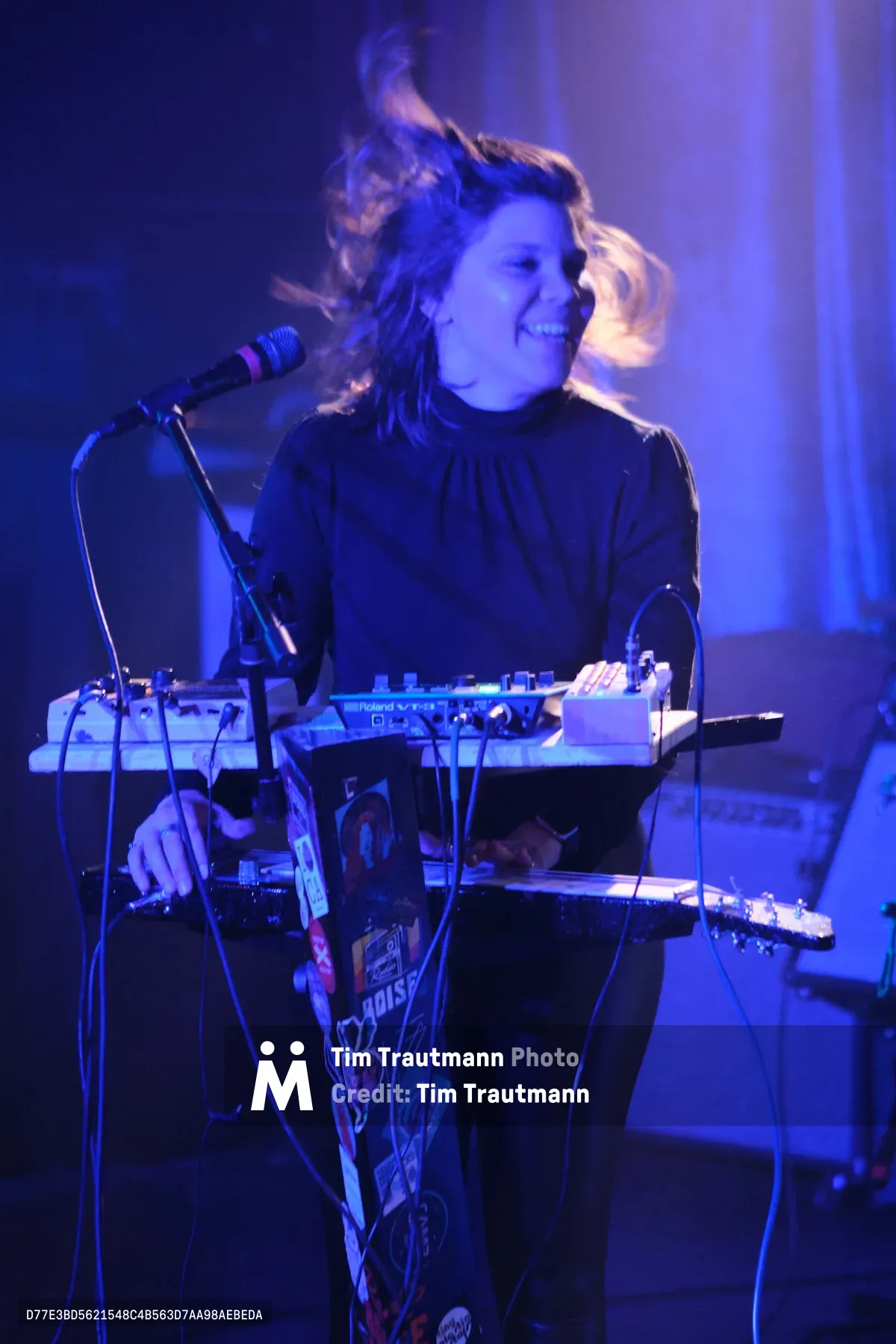A female electronic musician from experimental band Sun Blood Stories performs behind a Roland synthesizer setup at Dante's venue in downtown Portland, her blonde hair catching movement as she leans toward the microphone. Saturated blue and purple stage lighting bathes the intimate performance space, creating an otherworldly atmosphere that transforms the musician and her equipment into silhouettes against vertical light columns. The industrial venue's brick walls dissolve into darkness beyond the concentrated beam lighting, while cables snake across the stage connecting her multi-tiered keyboard rig.