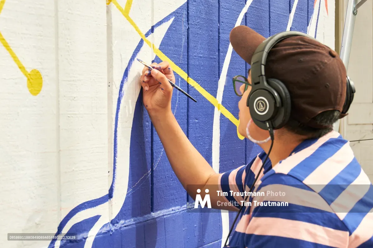 Maria Rodriguez, known as Sparkykneecap, works intently on her vibrant mural "Let's Talk" at Open Signal in Portland's Eliot neighborhood. Wearing headphones and a blue-striped shirt, the Mexican-American artist carefully applies precise brushstrokes to the bold geometric design featuring brilliant blues and yellows against white brick. The concentrated creative process unfolds in natural daylight, capturing the meditative focus required for large-scale public art.