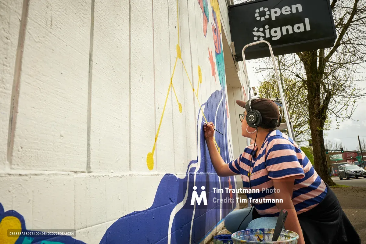 Maria Rodriguez, known as Sparkykneecap, applies vibrant blue paint to a large-scale mural on the exterior wall of Open Signal in Portland's Eliot neighborhood. Working from a ladder beneath the organization's black signage, the Mexican-American artist wears headphones and a striped shirt while adding intricate details to the commissioned piece titled 'Let's Talk.' The white corrugated metal surface comes alive with bold geometric forms and flowing colors that reflect Rodriguez's exploration of identity and cultural themes through playful, accessible art.