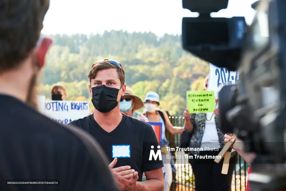 Michael Gaskill, wearing a black face mask and v-neck shirt, stands composed as he speaks to a KOIN Channel 6 news reporter during a climate change demonstration outside Representative Kurt Schrader's office in Oregon City. Behind him, fellow protesters hold signs including "Climate Crisis Is NOW" and "VOTING RIGHTS" against the backdrop of Oregon's rolling forested hills. The scene captures the intersection of grassroots activism and media coverage in the afternoon light of McLoughlin Promenade.