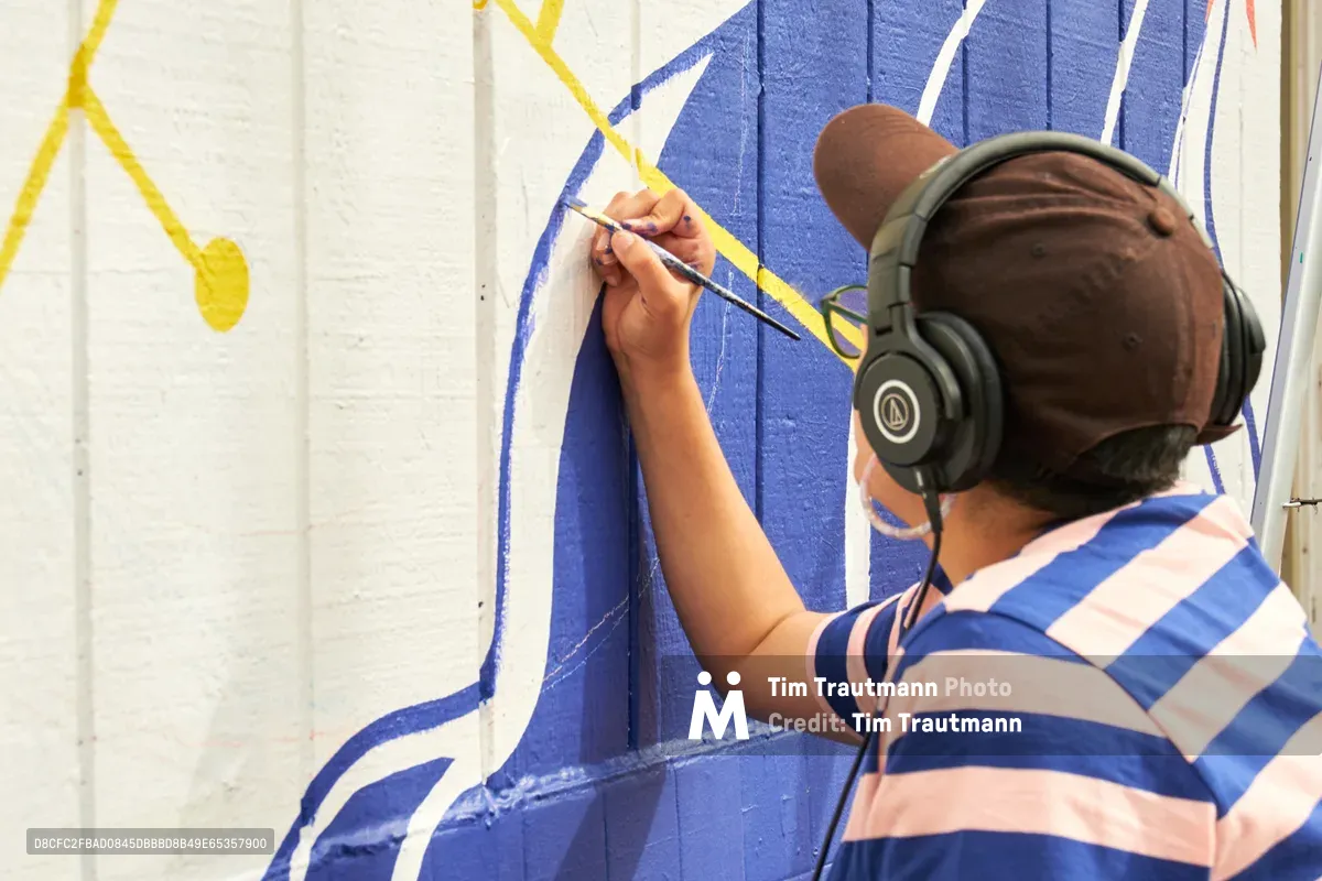 Mexican-American artist Maria Rodriguez, known as Sparkykneecap, meticulously details a vibrant blue and yellow mural at Open Signal in Portland's Eliot neighborhood. Wearing black headphones and a blue-striped shirt, they work with focused concentration on the community-commissioned piece titled 'Let's Talk.' The afternoon light illuminates their precise brushwork against the bold geometric patterns that explore themes of identity and cultural connection.