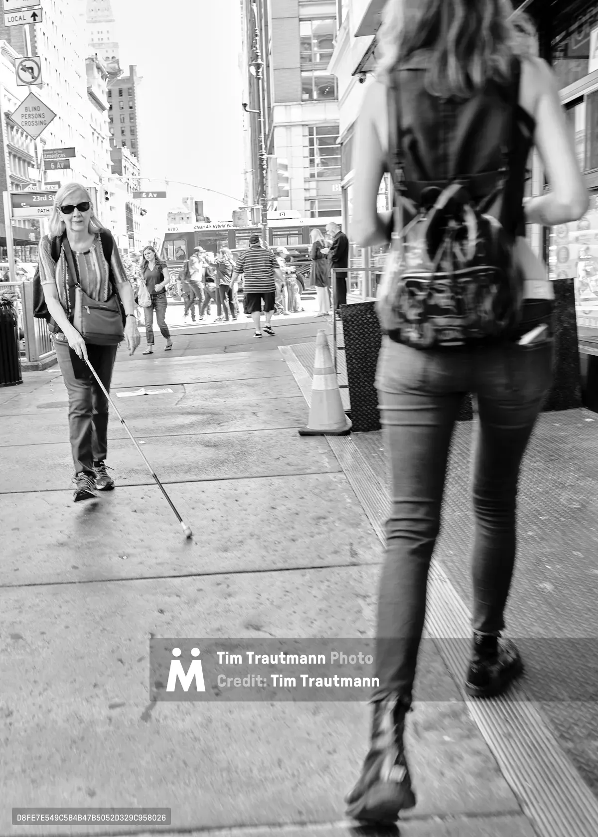 A striking black and white street photograph captures two figures moving in opposite directions along the bustling sidewalks of Chelsea's West 23rd Street. In the sharp foreground, a woman with flowing hair strides purposefully past the camera, while in the softly focused background, a blonde woman wearing sunglasses and carrying a white cane navigates the urban landscape with quiet determination. The dramatic contrast between the crystal-clear foreground subject and the deliberately blurred background creates a powerful visual metaphor for the different ways we all move through the city's relentless rhythm.