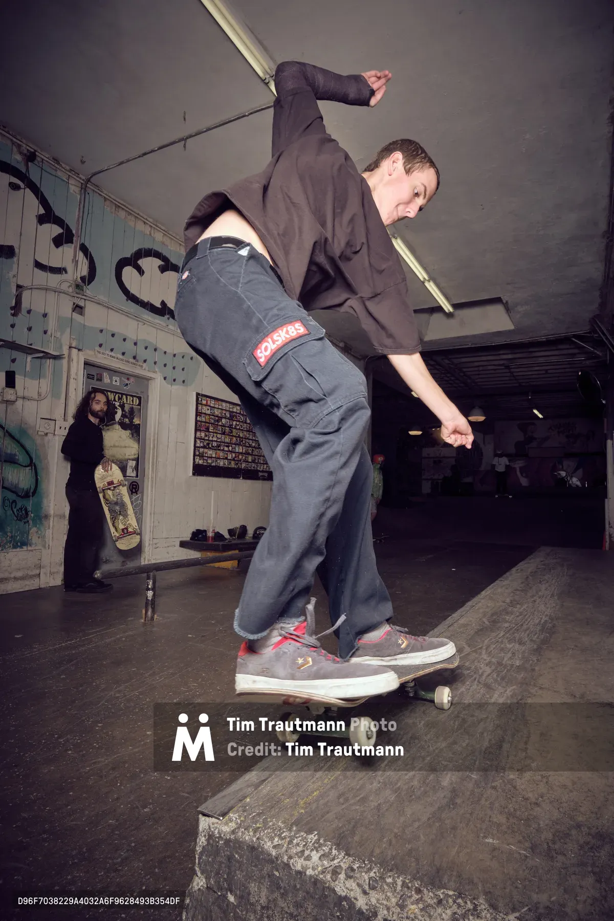 A skateboarder in dark casual wear and gray pants carves through the weathered concrete transitions of Portland's Commonwealth Skateboarding shop. The industrial interior buzzes with underground energy, graffitied walls and skateboard memorabilia creating an authentic backdrop. Fluorescent lighting casts dramatic shadows as the rider flows through the bowl with focused intensity, embodying the raw spirit of Pacific Northwest skate culture.
