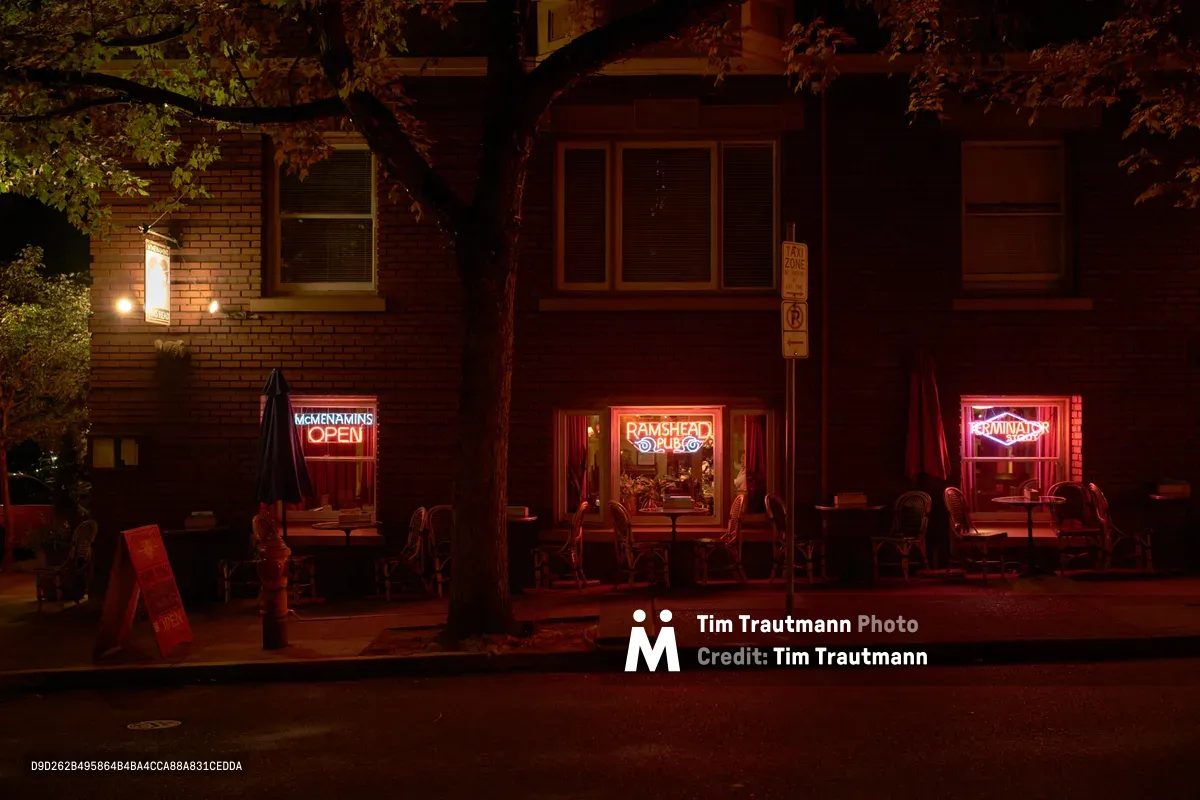 Crimson neon signs pierce the twilight darkness at McMenamins Ram's Head Pub in Portland's Alphabet Historic District, casting an inviting ruby glow across empty sidewalk tables and chairs. The historic brick building's warmth contrasts with the cool shadows of overhanging maple branches, while golden interior light spills through tall windows onto Northwest Hoyt Street. A solitary parking meter stands sentinel beside the welcoming entrance, marking this corner of Nob Hill's evening quiet.