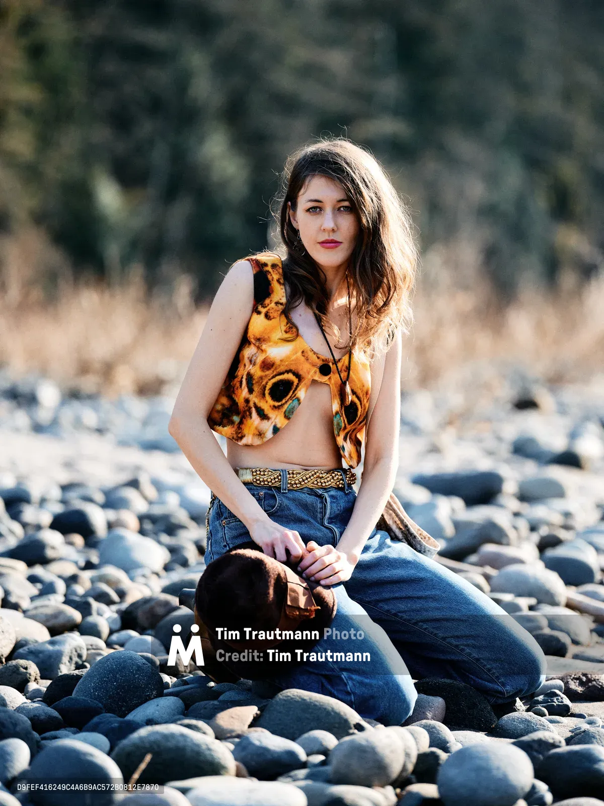 A young woman with flowing brown hair kneels gracefully among smooth river stones along Oregon's Floodplain Trail in Multnomah County. She wears a vibrant orange patterned crop top and denim jeans cinched with a woven belt, her pose contemplative against the sun-dappled backdrop of golden grassland and distant forest. The warm afternoon light creates a natural rim lighting effect, highlighting her figure against the softly blurred woodland setting.