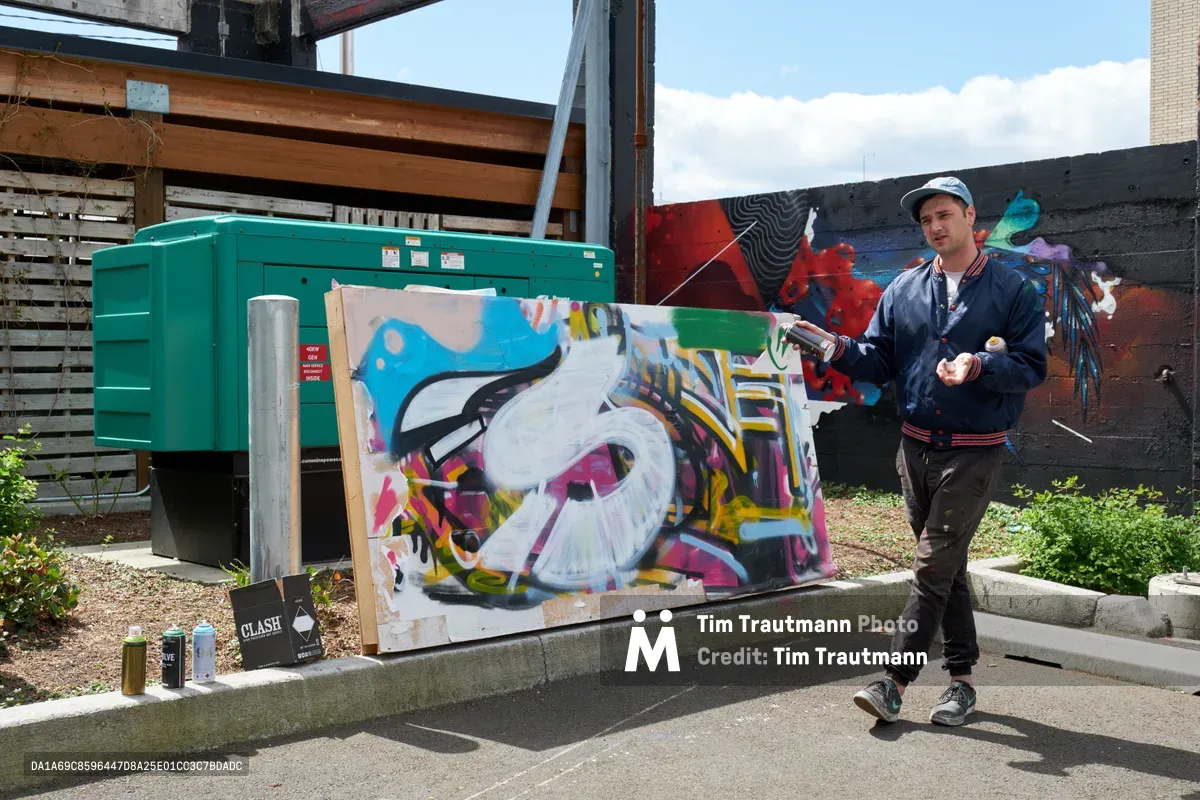 Galen Malcolm of the Portland Street Art Alliance holds a spray can while standing beside his vibrant graffiti artwork in progress at the Taylor Electric Blocks in Portland's Hosford-Abernethy neighborhood. The dynamic composition captures the artist mid-demonstration, with colorful abstract letterforms sprawling across a concrete wall behind him, while industrial green dumpsters and urban infrastructure frame the creative scene. Afternoon light illuminates the working artist's studio jacket and baseball cap as he engages with viewers during this educational street art tour.