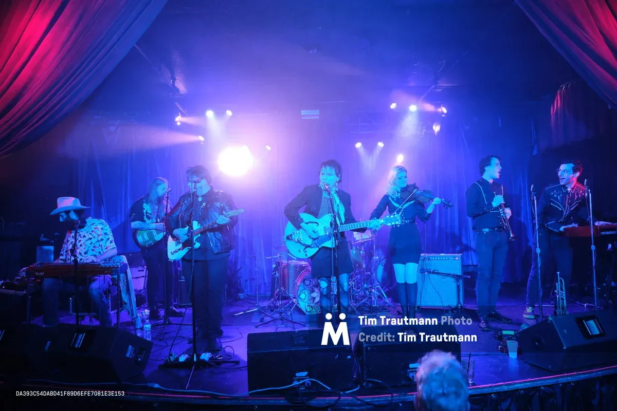 Seven musicians command the intimate stage at Dante's in Portland, bathed in electric blue stage lighting that cuts through veils of atmospheric haze. The band Roselit Bone performs beneath dramatic crimson curtains that frame the venue's signature cabaret atmosphere, their silhouettes emerging from the smoky darkness as spotlights pierce the ethereal performance space. Guitars, violin, and keyboards blend in this underground music sanctuary where Portland's alternative scene converges.