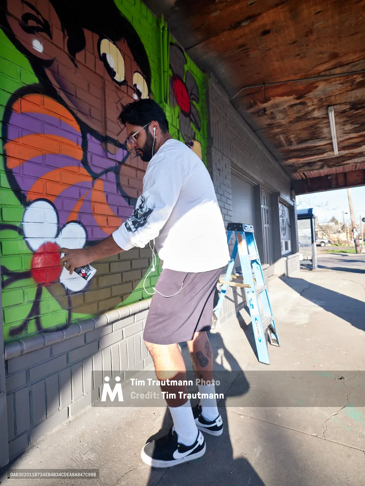 Muralist Heysus works intently on a colorful graffiti-style mural featuring cartoon faces and bold lettering on a brick wall in Portland's Alberta district. Dressed casually in a white sweatshirt and purple shorts, the bearded artist holds a spray can while standing on the concrete sidewalk beneath a weathered wooden overhang. The vivid green, orange, and red artwork transforms the urban storefront, with a stepladder positioned nearby and bright afternoon sunlight casting sharp shadows across the scene.