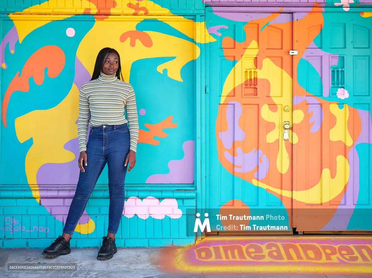 Artist Naomi Likayi stands beside her completed mural on Northeast 15th Avenue in Portland's Alberta district, her hand touching the vibrant wall art she has just signed. The explosive composition features flowing organic shapes in turquoise, coral, yellow, and purple that transform the building's facade into a celebration of color and movement. Wearing a striped turtleneck and dark jeans, Likayi poses confidently against her creation, which incorporates whimsical elements like floating clouds and abstract forms that seem to dance across the architectural surface.