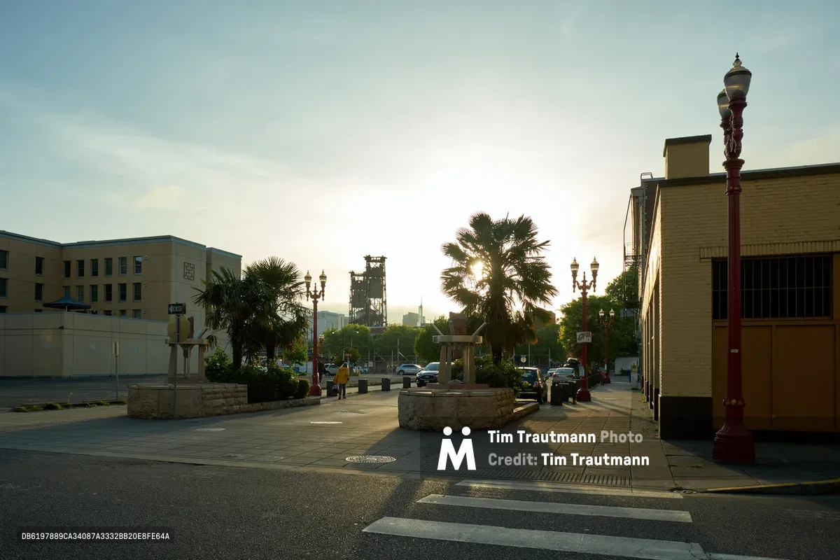The sun rises behind palm trees in Portland's Chinatown-Japantown Historic District, casting warm light across Northwest Everett Street. Ornate red street lamps stand sentinel over empty sidewalks and parked cars, while mid-century buildings frame the scene. A circular stone planter anchors the foreground as the industrial skyline dissolves into the hazy morning atmosphere.