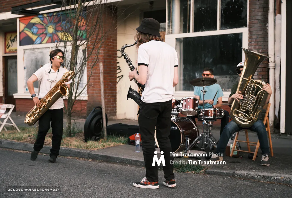 Four musicians animate the sidewalk during Portland's Last Thursday street fair on Northeast Alberta Street, their brass instruments catching the golden afternoon light. A saxophone player in sunglasses stands prominently on the left while a bassist in a black bucket hat commands center stage, flanked by a drummer and tuba player seated on folding chairs. The urban backdrop features weathered brick buildings adorned with colorful murals, creating an authentic Portland street scene where community and creativity converge.