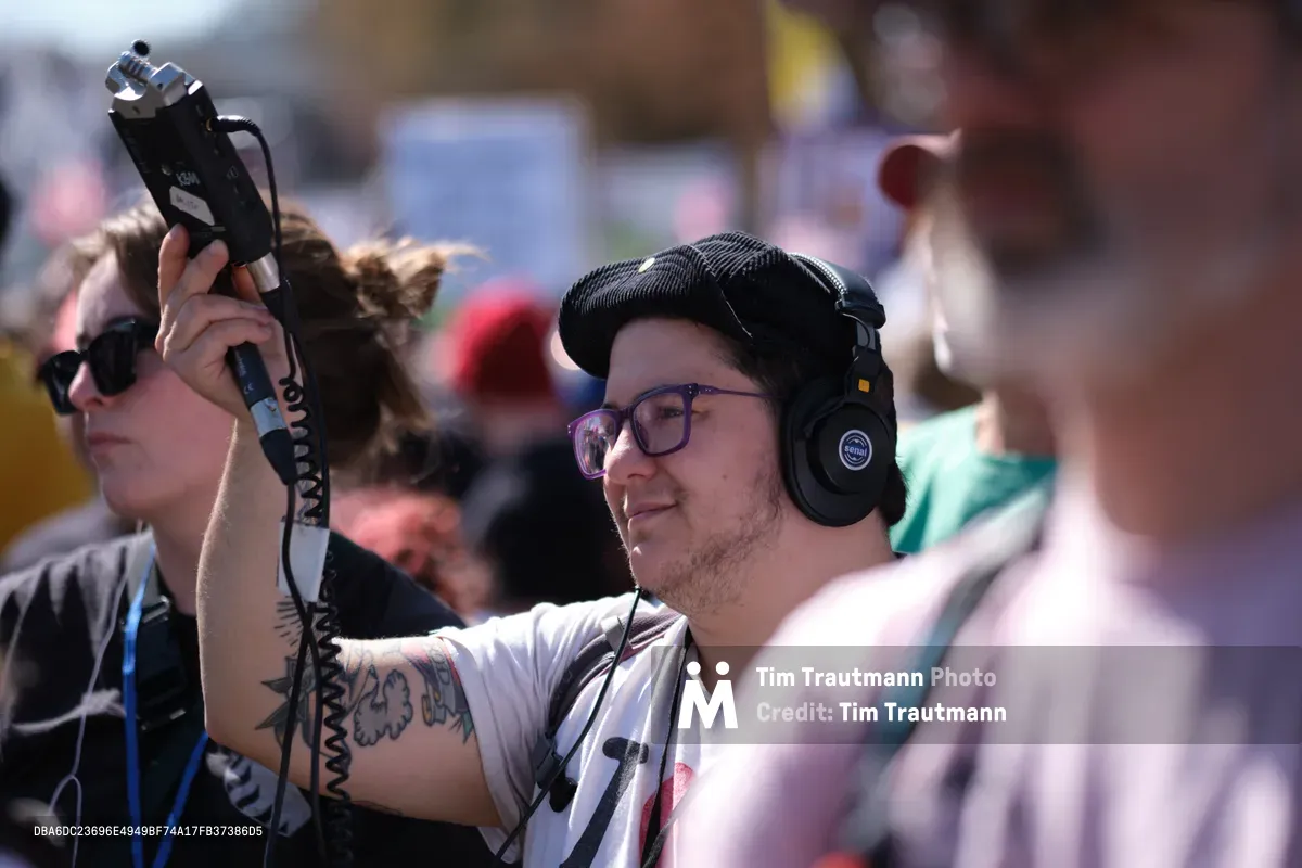 A focused sound technician wearing professional headphones and glasses captures audio at a protest gathering in Portland's historic Skidmore district. Golden afternoon light illuminates his concentrated expression as he operates recording equipment, while a tattooed colleague holds a boom microphone overhead. The warm sunlight creates a striking contrast against the urban backdrop, highlighting the dedicated media professionals documenting this March 2026 anti-monarchy demonstration.
