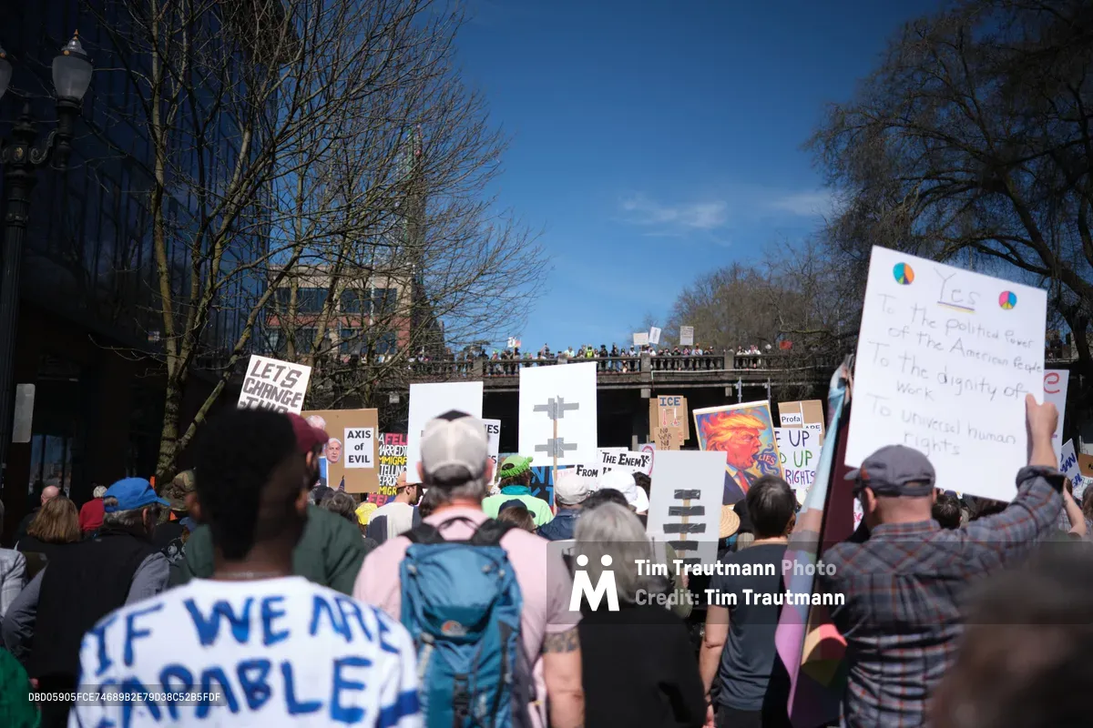 Demonstrators gather beneath Portland's historic Burnside Bridge in the Old Town district, their handmade signs creating a forest of dissent against azure spring skies. The crowd flows along Southwest Ankeny Street, with protesters of all ages holding placards declaring "LET'S CHANGE" and "IF WE ARE CAPABLE" among other messages of resistance. Bare-branched trees frame the scene while onlookers line the bridge above, witnessing this grassroots political mobilization in the heart of Portland's Chinatown neighborhood.