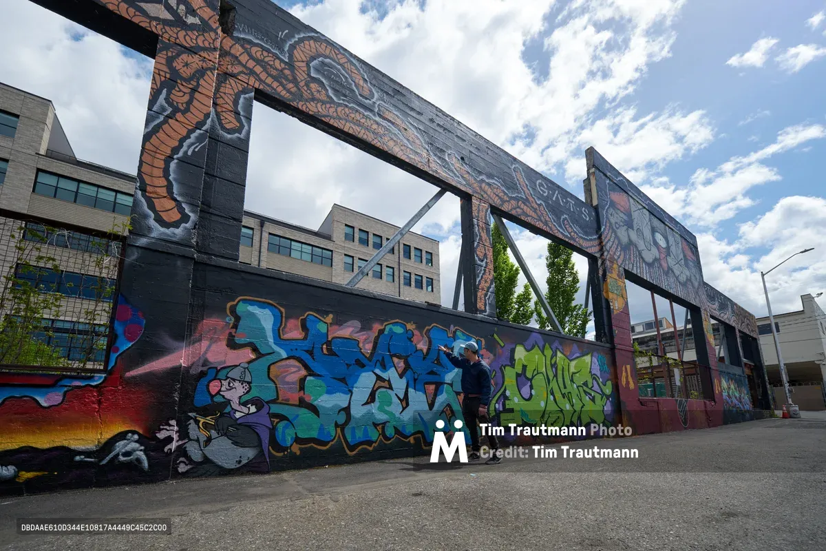 A guide from the Portland Street Art Alliance leads an interpretive tour beneath a towering industrial bridge adorned with vibrant graffiti murals in Portland's Hosford-Abernethy neighborhood. The scene captures the intersection of urban decay and creative renewal, where colorful street art transforms concrete infrastructure into an outdoor gallery. Dramatic clouds frame the composition, highlighting the contrast between the gritty industrial setting and the dynamic artistic expression that covers every available surface.