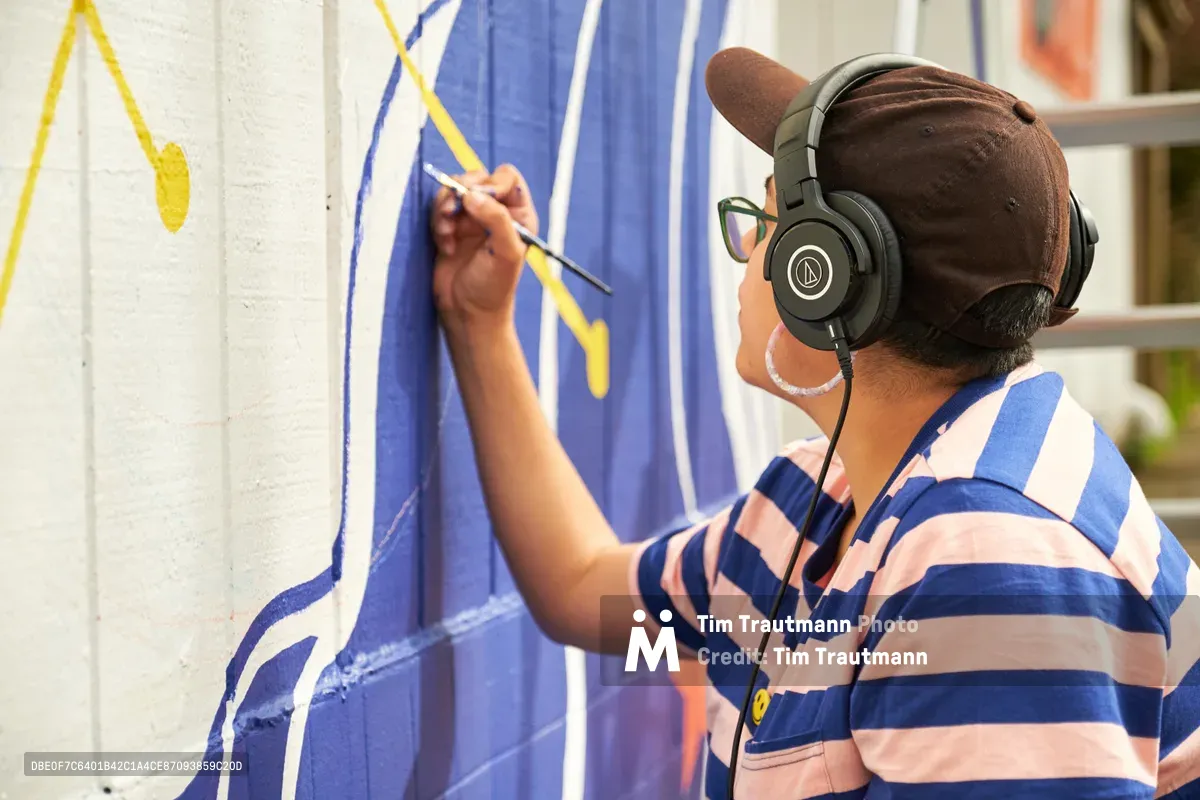 Mexican-American artist Maria Rodriguez, known as Sparkykneecap, works intently on her vibrant blue and yellow mural titled "Let's Talk" at Open Signal in Portland's Eliot neighborhood. Wearing headphones and a brown cap, she applies precise brushstrokes to the wall while dressed in a blue and white striped shirt. The afternoon light illuminates her focused expression as she brings themes of identity and cultural connection to life through bold geometric forms and warm colors.