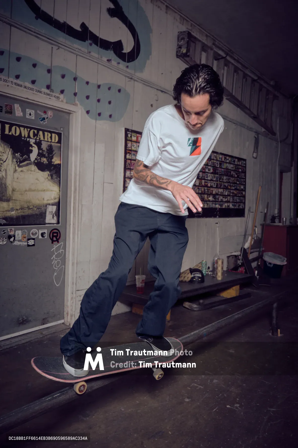 A concentrated skater navigates his board through  Commonwealth Skateboarding in Portland's Buckman neighborhood. Dramatic overhead lighting cuts through the dim space, illuminating vintage skate posters and memorabilia that plaster the weathered walls. The intimate setting captures the raw authenticity of underground skate culture, where passion thrives in unconventional spaces.