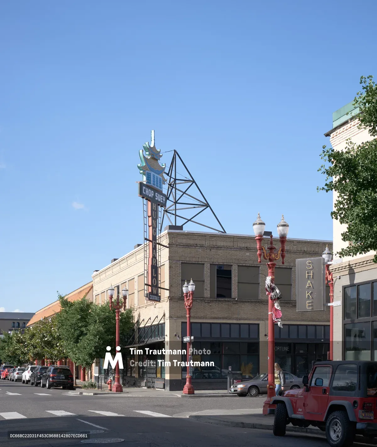 A weathered vintage Chop Suey neon sign towers above the brick storefronts of Portland's historic Chinatown district, its geometric metal framework stark against the clear blue afternoon sky. The iconic pagoda-topped sign anchors a streetscape of early 20th-century commercial buildings, where red decorative lampposts and parked vehicles line the quiet urban thoroughfare. Dappled sunlight filters through mature street trees, casting gentle shadows across the pavement and highlighting the architectural details of this culturally significant neighborhood.