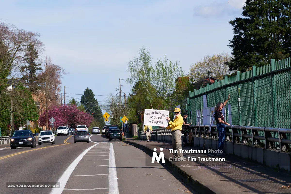 Two activists stand on the North Skidmore Street overpass in Portland's Humboldt neighborhood, displaying a protest banner reading 'No Work No School No Shopping May 1st' to passing traffic on Interstate 5 below. The scene unfolds under a soft spring sky, with one demonstrator in a bright yellow jacket holding the white banner while another figure stands nearby. Vehicles flow beneath the green-fenced pedestrian bridge as blooming trees frame this moment of grassroots political action.