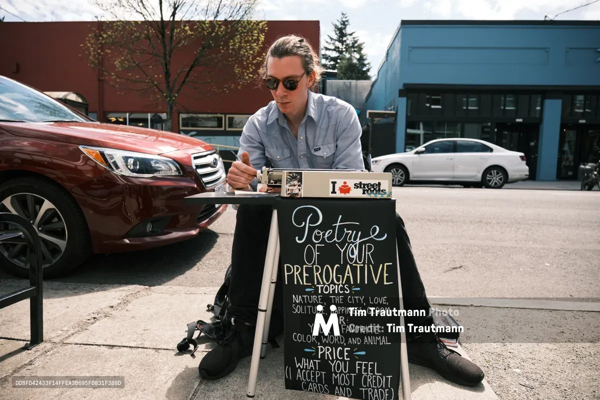 Till Gwinn, a local street poet, sits with legs spread apart on the sidewalk along Northeast Alberta Street in Portland, Oregon, crafting personalized poetry for passersby. Dressed in a light blue button-down shirt and dark sunglasses, he works intently at a small folding table adorned with a chalkboard sign advertising "Poetry of Your Prerogative Topics." The urban scene unfolds behind him with parked cars, brick buildings, and the characteristic overcast Pacific Northwest sky filtering soft, even light across the pavement.