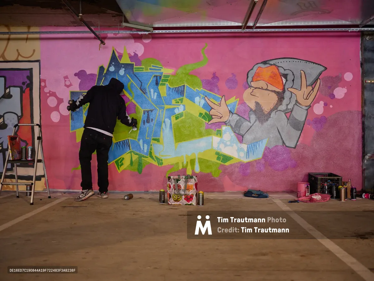 A black-clad artist works intently on a vibrant mural featuring dimensional blue and green lettering alongside a bearded character throwing up peace signs, all set against a hot pink backdrop in Clay Creative's parking garage. The industrial space comes alive with creative energy as spray paint cans and art supplies scatter across the concrete floor. Exposed ceiling elements and raw concrete walls frame this underground artistic sanctuary in Southeast Portland's Hosford-Abernethy neighborhood.
