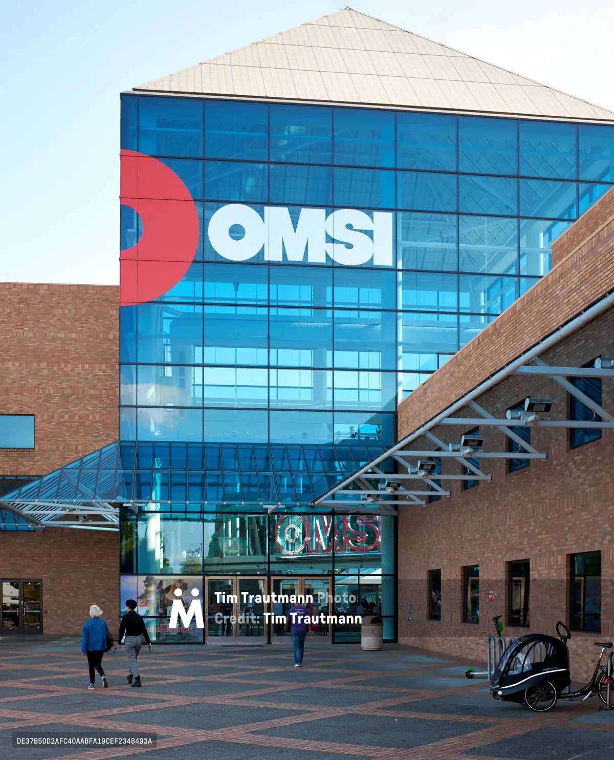 The Oregon Museum of Science and Industry's striking contemporary facade dominates this evening view, where warm brick construction contrasts dramatically with expansive blue-tinted glass panels bearing the iconic OMSI logo. Visitors move leisurely across the geometric brick plaza as twilight approaches, while modern steel canopies frame the museum's main entrance. The architectural interplay of transparent and solid surfaces creates a dynamic composition that captures the institution's blend of accessibility and innovation along Portland's industrial waterfront.