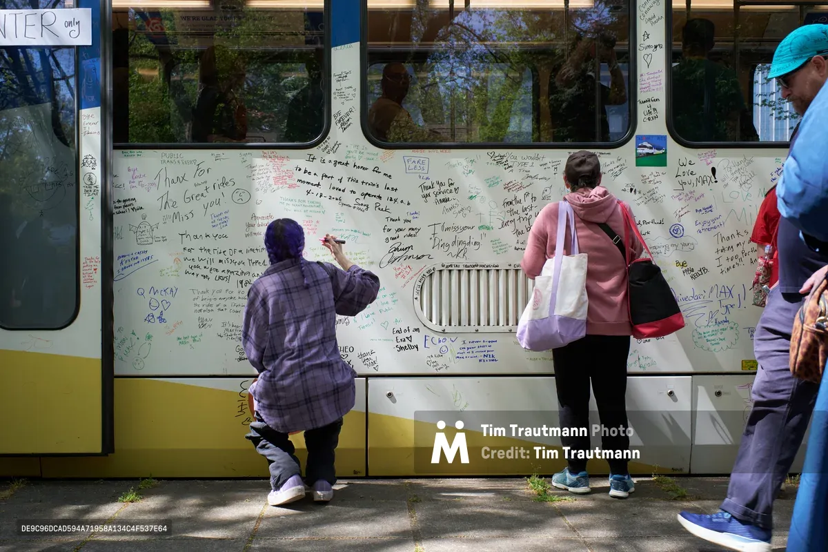 Commuters and transit enthusiasts gather around a retiring TriMet MAX Type 1 light rail car in Portland's Lloyd District, adding their handwritten messages and signatures to the vehicle's white exterior. A person in a plaid shirt crouches while writing on the train's side as others stand nearby with bags and belongings, creating an intimate farewell scene bathed in dappled afternoon sunlight. The train's windows reflect the surrounding urban trees, while countless personal messages in various inks cover nearly every inch of the accessible surface.