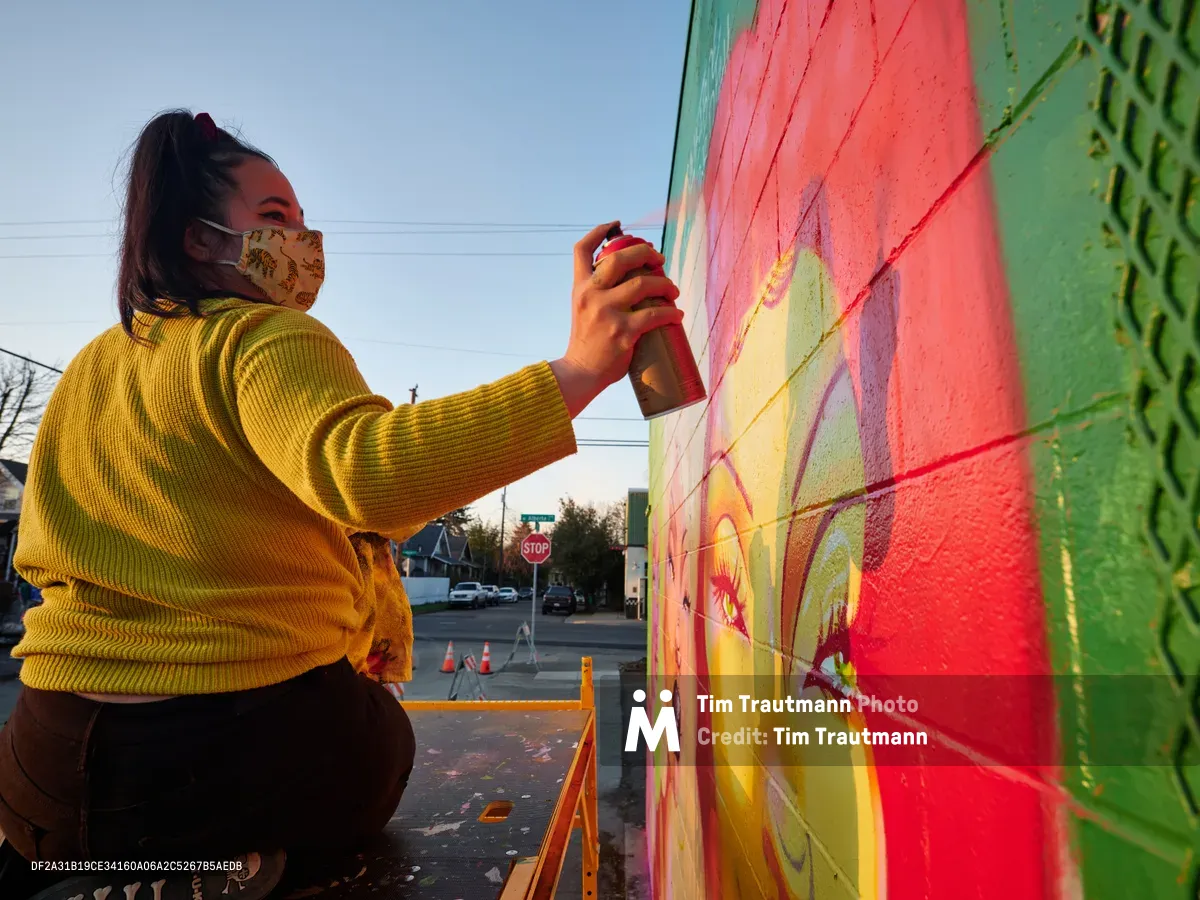 Multidisciplinary artist Vilatose, wearing a golden corduroy sweater and patterned face mask, spray paints bold coral and lime green hues onto her mural 'Jardín Cubana' featuring three female figures on a wall in Portland's Alberta Arts District. The late afternoon light bathes the scene in warm tones as she works methodically on the colorful mural, with urban street elements and traffic cones visible in the background. The composition captures the intimate moment of artistic creation against the backdrop of Northeast Portland's creative corridor.