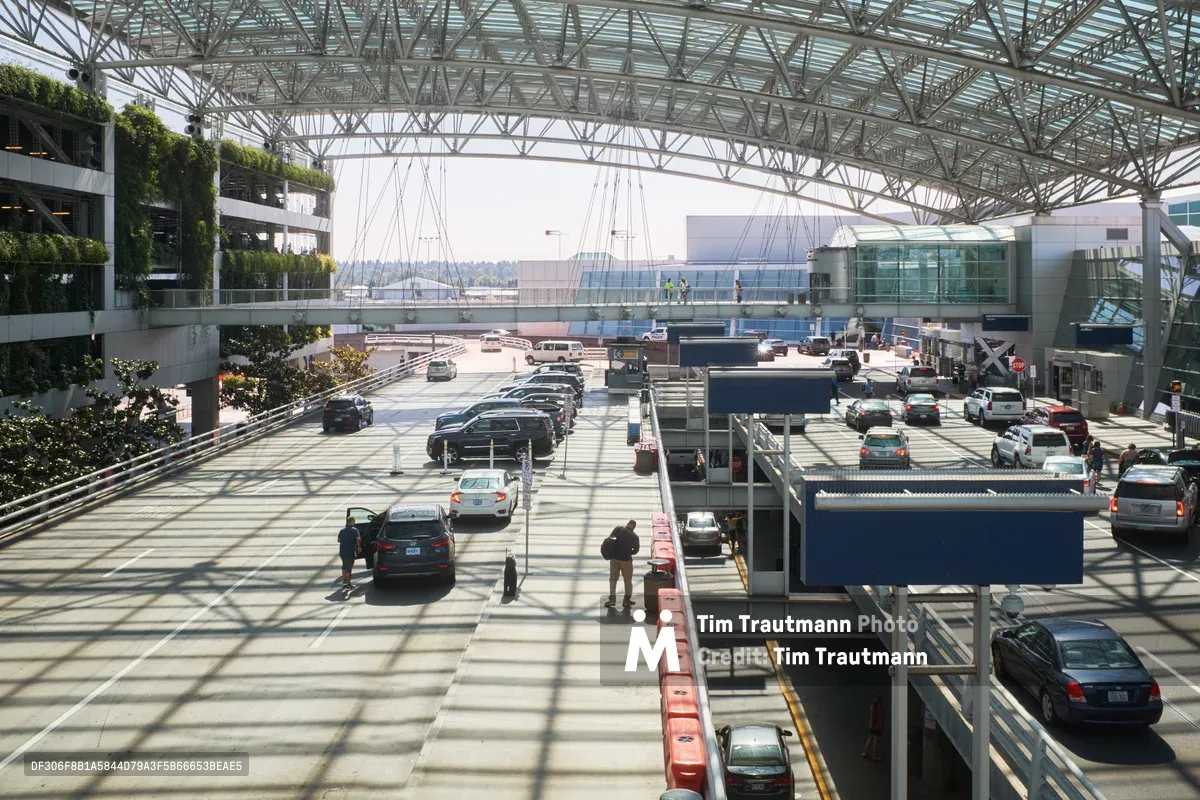 The multi-level departures roadway at Portland International Airport bustles with vehicular activity beneath a soaring canopy of steel trusses and glass. Cars queue in organized lanes while passengers load luggage and navigate the concrete plaza, creating a choreographed dance of arrival and departure. Dappled shadows from the architectural framework create geometric patterns across the pavement, while the terminal's modern glass facade gleams in the background. The scene captures the perpetual motion of modern air travel infrastructure, where engineering precision meets human transit needs.