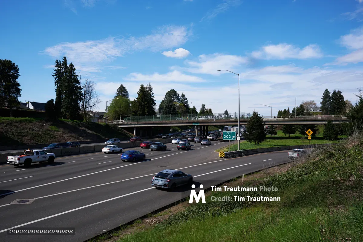 Activists from the Indivisible movement stage a banner drop from a pedestrian overpass spanning Interstate 5 in North Portland, their protest banner visible against the spring sky as afternoon traffic flows beneath. The concrete bridge arcs over multiple lanes of busy freeway traffic, with evergreen trees and residential neighborhoods framing the scene under wispy clouds. This moment captures civic engagement intersecting with daily commuter life, as the activists' May 1st General Strike message reaches drivers below on one of the Pacific Northwest's major transportation arteries.