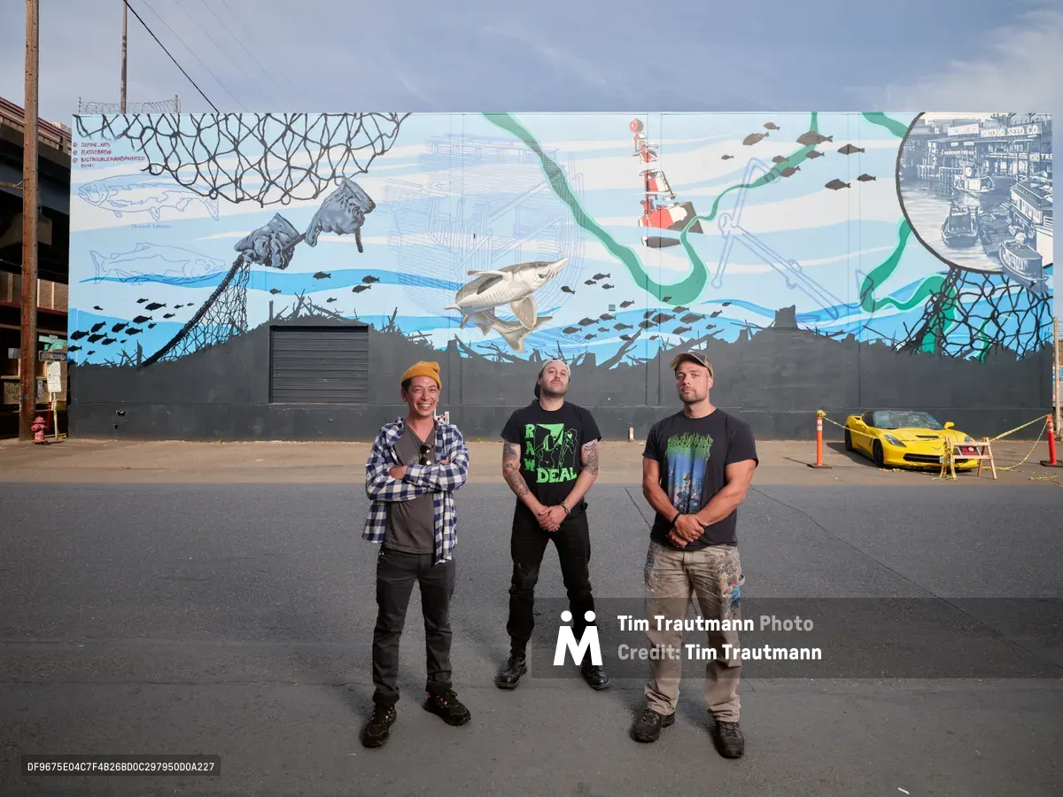 Three muralists stand proudly before their completed marine-themed artwork on a concrete wall in Southeast Portland's Central Eastside district. The vibrant mural depicts an underwater ecosystem with whales, fish, and maritime vessels rendered in blues and greens, while the artists - Jeremy Nichols in flannel and beanie, bigtroublehandpainted in black attire, and Devin Finley in work clothes - pose confidently on the industrial street. A yellow sports car and urban infrastructure frame the scene under an overcast Pacific Northwest sky.