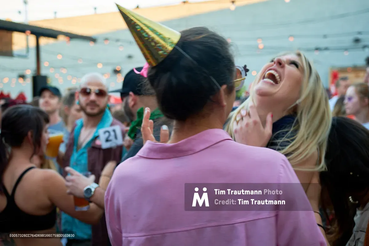 A blonde woman throws her head back in unrestrained laughter during Portland's Your Sunday Best day party at White Owl Social Club. The golden party hat catches the warm afternoon light as string lights create bokeh in the background. A man in a soft pink shirt leans in beside her, while fellow partygoers with drinks populate the festive outdoor scene.