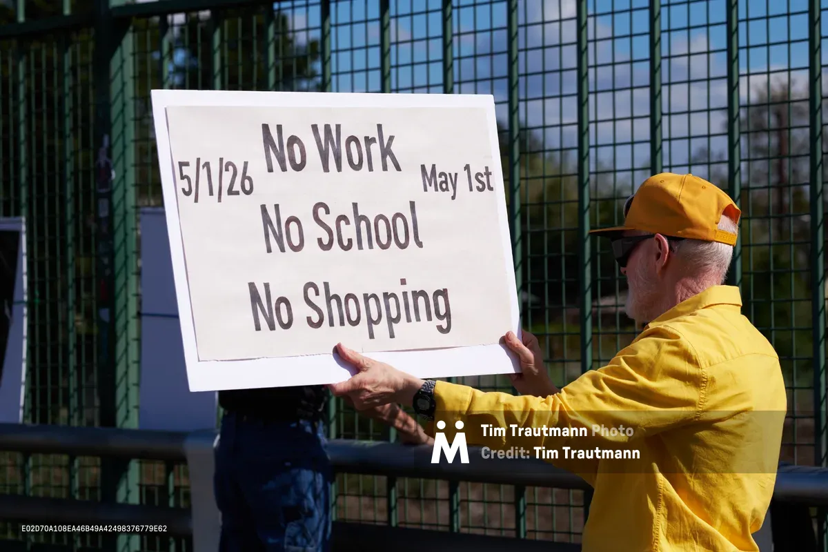 An activist in a vibrant yellow windbreaker and matching cap holds aloft a handwritten protest banner over Interstate 5 in North Portland's Skidmore neighborhood. The stark white sign bearing "5/1/26 No Work No School No Shopping May 1st" catches afternoon sunlight against the geometric shadows of a chain-link overpass fence. Industrial architecture frames this moment of grassroots organizing, as the demonstrator's weathered hands grip the edges of their call to action.