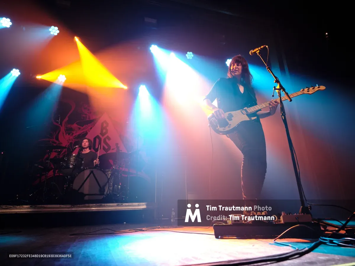 Emma Richardson commands the stage with her white bass guitar while Julian Dorio anchors the rhythm from behind his drum kit during Band of Skulls' 10th anniversary tour at Webster Hall in Manhattan. Dramatic stage lighting cuts through theatrical smoke, casting amber and cyan beams that illuminate the duo against the venue's shadowy backdrop. The intimate concert setting captures the raw energy of their milestone celebration, with Richardson positioned prominently in the foreground as colored spotlights create an atmospheric haze around the performance space.