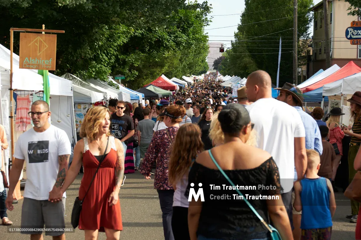 A vibrant street festival transforms North Mississippi Avenue in Portland, Oregon, with hundreds of visitors flowing between white vendor tents that line the closed road. The afternoon sun filters through mature street trees as families, couples, and individuals of all ages browse local artisans and food vendors. A diverse crowd moves leisurely through the pedestrian-only space, creating an atmosphere of community celebration and local commerce.