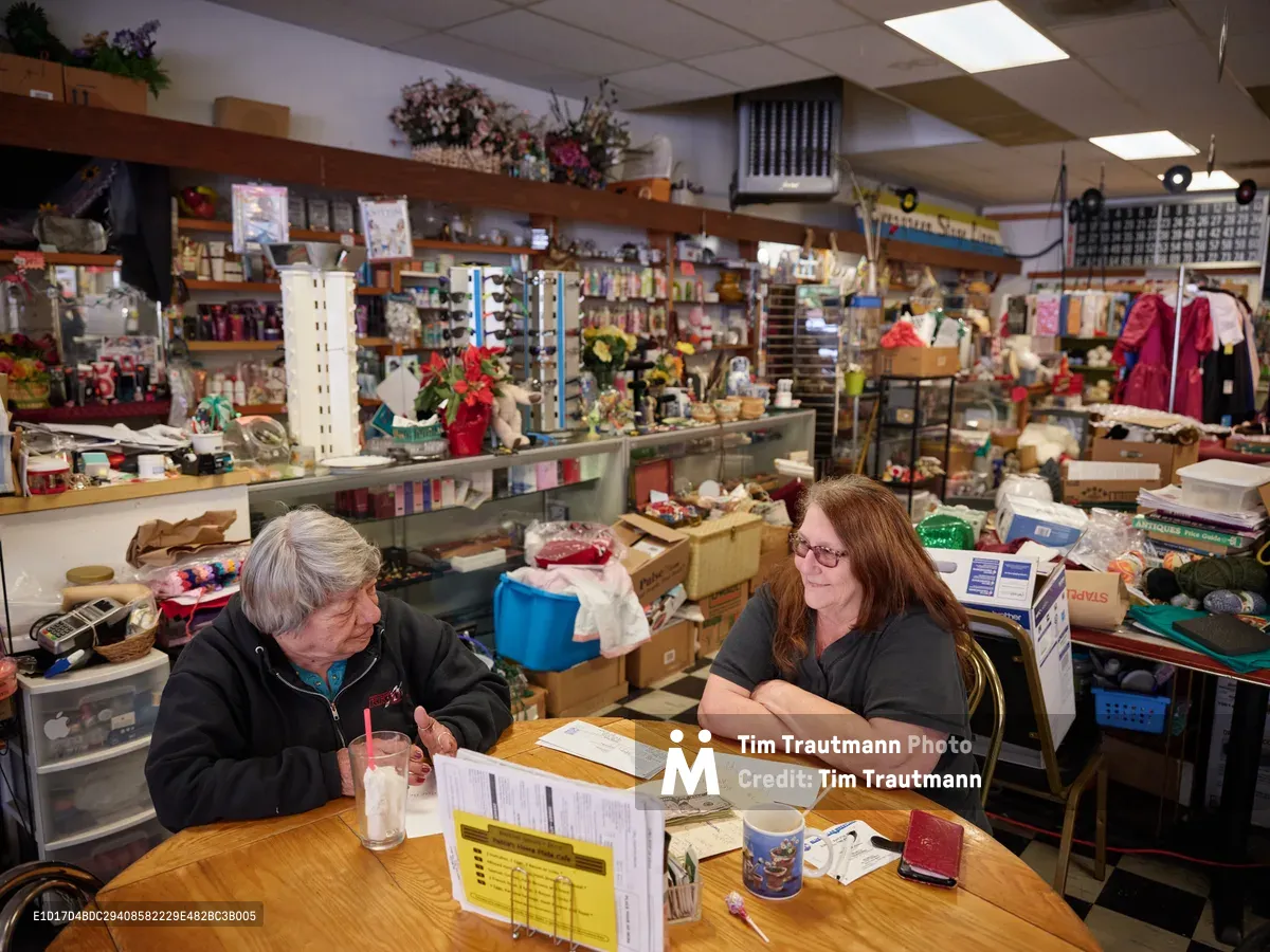 Pattie Deitz, owner of Pattie's Home Plate Café in Portland's Saint Johns neighborhood, shares an intimate moment with her best friend Jenny across a worn wooden table scattered with papers and coffee cups. The fluorescent-lit interior reveals a treasure trove of craft supplies, vintage collectibles, and homewares that transform this North Lombard Street establishment into more than just a restaurant. Their genuine connection radiates warmth against the backdrop of organized chaos that defines this beloved community gathering place.