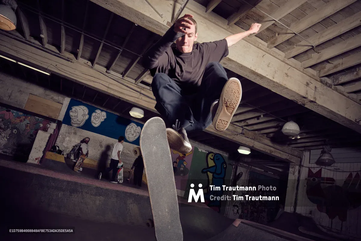 A skateboarder suspended mid-air executes a dynamic trick in the atmospheric basement of Commonwealth Skateboarding in Portland's Buckman district. The concrete space pulses with underground energy, its exposed wooden ceiling beams and colorful graffitied walls creating a raw, authentic backdrop. Dramatic lighting cuts through the subterranean setting, illuminating the athlete's focused expression and the worn grip tape of his deck as he floats momentarily above the industrial floor.