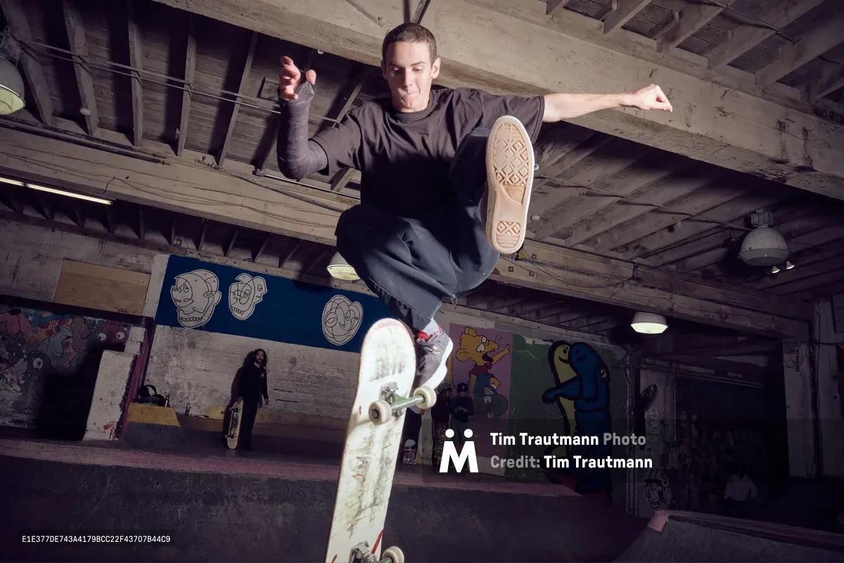 A skateboarder launches into a dynamic aerial maneuver inside Commonwealth Skateboarding in Portland's Buckman neighborhood. The raw concrete and exposed wooden beams of the indoor skate space frame the athlete mid-flight, while colorful skull artwork and graffiti murals create a gritty urban backdrop. The dramatic low-angle perspective captures the precise moment of weightless suspension, with the skater's weathered deck clearly visible beneath his sneakers.