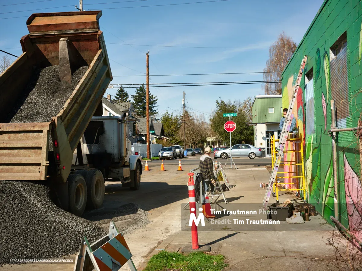 Artist Villatose works from a ladder against the vibrant green wall of a building on Northeast Alberta Street in Portland's Alberta Arts District, creating the mural titled Jardín Cubana featuring three female figures. A dump truck loaded with gravel dominates the left foreground while orange traffic cones mark the construction zone on this bright afternoon. The scene captures the intersection of infrastructure development and creative expression that defines this evolving neighborhood.
